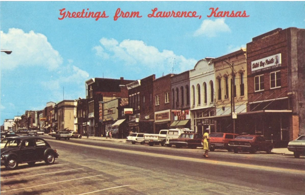 Vintage postcard of downtown Lawrence, Kansas, reading "Greetings from Lawrence, Kansas" in red script above a Massachusetts Street scene with mid-century cars parked along brick storefronts and a woman in a yellow dress crossing the street.